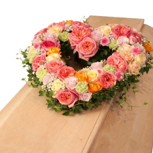 Hjärtformad blomsterkrans med rosor i rosa, orange och vita nyanser ligger ovanpå en kista av ljust trä.