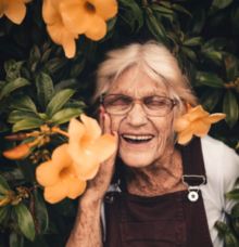 Äldre kvinna med glasögon ler omgiven av stora orange blommor och grönska.