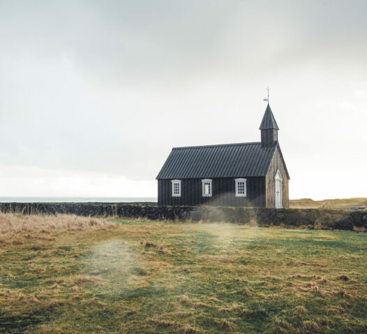 Liten svart kyrka med klocktorn ligger på en grässlätt under en molnig himmel.