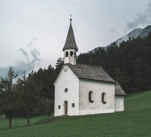 Vit kyrka med ett klocktorn står på en grönsluttning framför skog och berg.