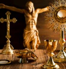 En samling religiösa föremål i guld, inklusive ett krucifix och en monstrans, står på ett bord framför en Jesusstaty.