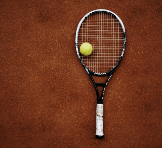 En tennisracket och en gul boll ligger på en grusplan.