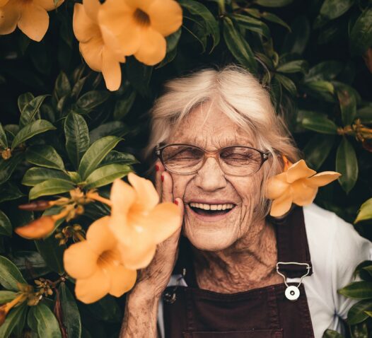 En äldre kvinna ler omgiven av gröna löv och orange blommor.